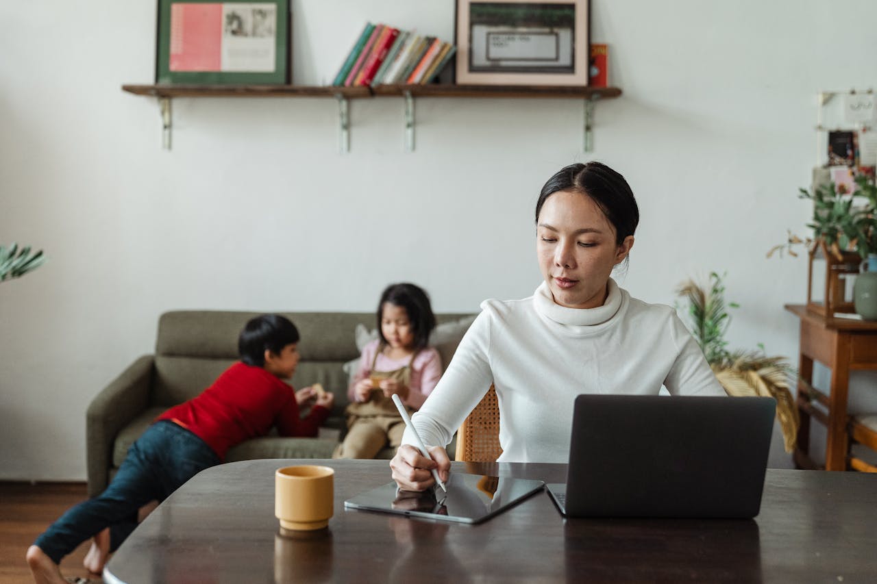 service-01 Mother multitasking with laptop and stylus alongside her children playing at home.